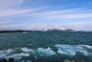 Buzdağları, arka planda karlı dağlar ve buzullarla kaplı İzlanda 'daki Jokulsarlon buzulu gölünün derin mavi sularında yüzüyor.