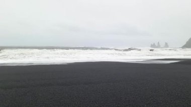 Reynisfjara 'ya vuran beyaz dalgalar arka planda bazalt deniz yığınları ve sisli uçurumlar olan siyah kum plajlarına çarpıyor.
