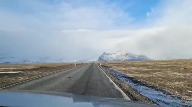 Karlı bir İzlanda manzarasında, arka planda dağlar olan asfalt bir yolda giden bir araç.