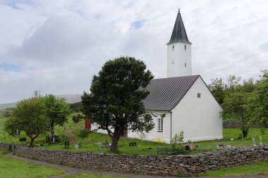 Holar, İzlanda, 12. Haziran 2024 'te, bulutlu bir günde İzlanda' nın Holar kentindeki bir mezarlığın üzerinde yükselen koyu gri çatılı ve yüksek çan kulesiyle beyaz köy kilisesi binası.