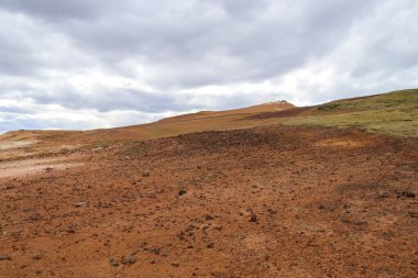 İzlanda 'da jeotermal bir bölgenin kırmızımsı yamaçları dramatik, bulutlu bir gökyüzünün altında seyrek bitki örtüsü sergiliyor ve eşsiz manzarayı vurguluyor.