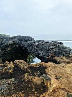 Okyanus dalgaları İzlanda 'nın engebeli kıyısında karamsar, bulutlu bir gökyüzü tarafından çerçevelenmiş dramatik bir volkanik kaya kemerine çarpıyor.