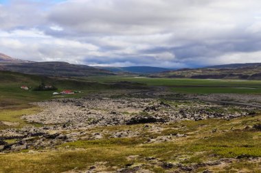 İzlanda 'daki Grabrok krateri yakınlarındaki manzarada bir lav tarlası, kırmızı çatılı çiftlik, dolambaçlı yol ve bulutlu bir gökyüzünün altındaki yeşil vadi yer alıyor.