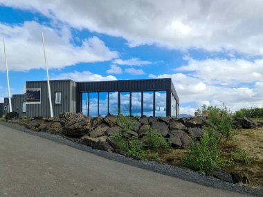 Skutustadahreppur, İzlanda 22. Haziran 2024, İzlanda 'daki mavi gökyüzünü ve bulutları yansıtan modern minimalist bina, kayalık bir duvar ve asfalt bir yol yakınında.