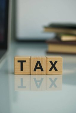 Vergi sözcüğünü gösteren üç tahta küp yansıtıcı bir yüzeye oturmuş, arka planda bulanık dosyalar, vergi sezonunu ve finansal planlamayı temsil ediyor.