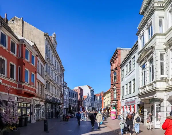 Flensburg, Almanya 'nın ana caddesinde yürüyen insanlar güneşli bir bahar gününde renkli binaların ve dükkanların keyfini çıkarıyorlar.
