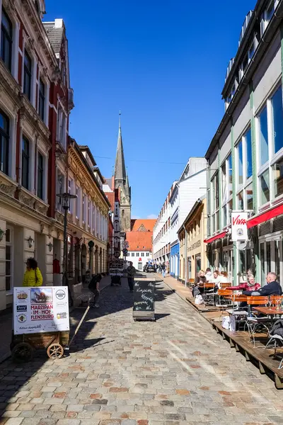 Flensburg, Almanya 'daki Cobblestone Caddesi' nde renkli binalar, açık kafeler ve St. Nikolai Kilisesi 'nin kulesi yer alıyor.