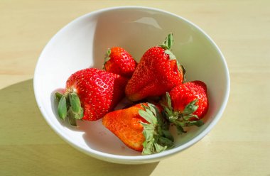 Olgun, yeşil yapraklı beş çilek, beyaz bir kasede, tahta bir yüzeyde, doğal ışıkla aydınlatılmış.