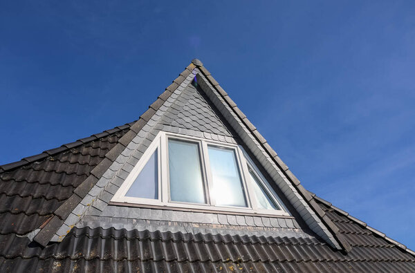 Треугольное окошко с белой рамой, отражающей солнечный свет на шиферной черепичной крыше жилого дома напротив ясного голубого неба