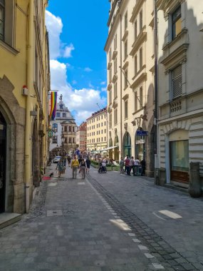 Münih, Almanya - 22. Haziran 2024: İnsanlar Münih şehir merkezinin yayalı bir caddesinde yürürken ve güneşli bir günün tadını çıkarırken, arka planda gökkuşağı bayrağı ve mavi gökyüzü görülüyor