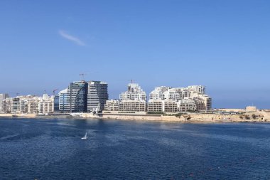 Valletta, Malta 28 Eylül 2024: Sliema kıyı şeridi boyunca uzanan modern binalar ve inşaat vinçleri açık mavi gökyüzünün altında, ön planda derin mavi Akdeniz suları ile