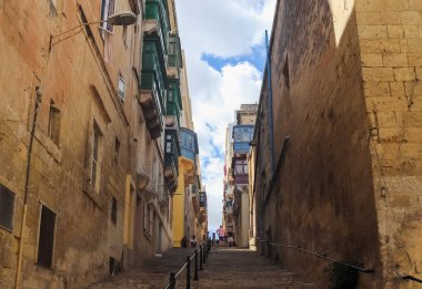 Valletta, Malta - 01. Ekim 2024: Turistler Valletta, Malta 'da renkli balkonlar ve tarihi kumtaşı binalarla çevrili dar bir sokakta yürüyorlar