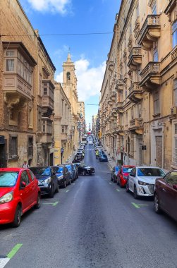 Valletta, Malta - 01. Ekim 2024: Valletta, Malta 'da dar bir sokakta mavi gökyüzünün altında arka planda bir kilise bulunan park edilmiş arabalar