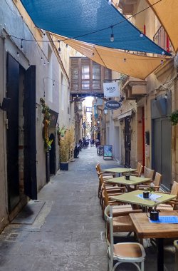 Valletta, Malta - 01. Ekim 2024: Valletta, Malta 'da güneşli gölgeler altında açık masa ve sandalyelerle büyüleyici dar bir cadde, akşam yemeği için hoş bir atmosfer yaratıyor.