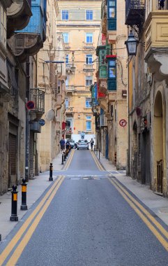 Valletta, Malta - 01. Ekim 2024: Valletta, Malta 'da renkli ahşap kaplı balkonları olan tipik dar cadde, asfalt yolda giden bir arabayı gösteriyor