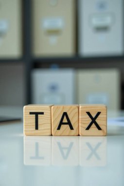 Vergi sözcüğü beyaz masa yüzeyine yansıyan tahtadan küplere, arka planda bulanık ofis dosyaları üzerine yazıldı