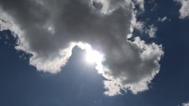 Parlak güneş ışığı koyu kümülüs bulutlarını delip gündüz vakti geniş mavi gökyüzü arkaplanına karşı dramatik atmosferik etki yaratır.