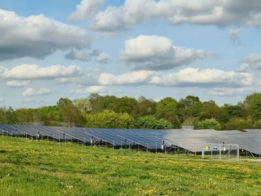 Güneş panelleri yeşil bir alanda duruyor. Parçalı bulutlu bir gökyüzünün altında temiz enerji üretiyorlar. Teknoloji ve doğanın uyumunu sergiliyorlar.