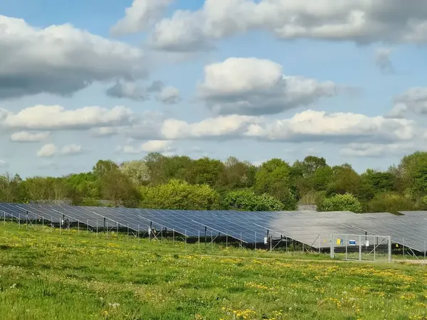 Güneş panelleri yeşil bir alanda duruyor. Parçalı bulutlu bir gökyüzünün altında temiz enerji üretiyorlar. Teknoloji ve doğanın uyumunu sergiliyorlar.