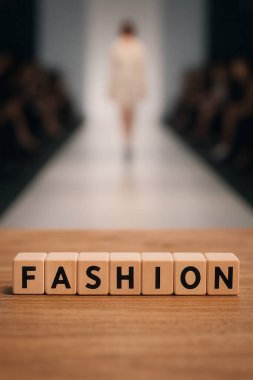 Moda sözcüğünü içeren ahşap küpler arka planda podyumda yürüyen bulanık bir model.