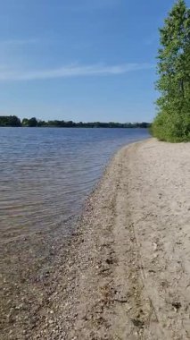 Sakin göl suyu, kumlu ve çakıllı bir kıyı şeridinde yavaşça süzülerek berrak mavi gökyüzünün altında küçük dalgalanmalar oluşturur.