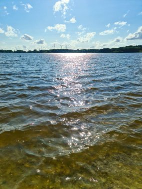 Güneş, arka planda rüzgâr çiftliği ve ormanı olan bir gölün hafif dalgalı su yüzeyine yansıyor. Mavi gökyüzünün altında bazı bulutlar var.