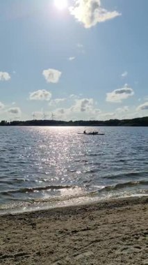 Kayakçılar güneşli bir günde kıyıdan uzakta kürek çekiyorlar. Arka planda bulutlar ve rüzgar türbinleri var.