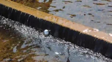 Yıpranmış, paslı fıskiyenin içine sıkışmış metal küre, pürüzsüz, parlak yüzeyi kısmen akan su akıntısına batmış kentsel manzarayı yansıtıyor.