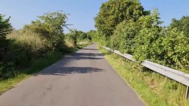 Çevresel asfalt yol yeşil kırsal arazide yayılıyor, uzak ufuk ile aydınlık yaz gökyüzü altında huzurlu kırsal araziyi ortaya çıkarıyor.