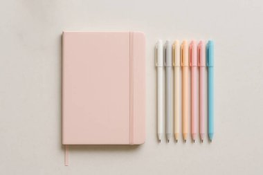 Bej bir yüzeye dizilmiş pembe defter ve pastel renkli kalemler not almak veya günlük tutmak için şık ve düzenli bir çalışma alanı oluşturur.