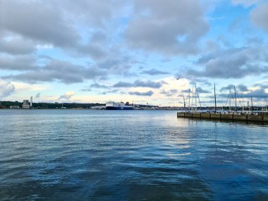 Kiel, Almanya - 02. Ağustos 2025: Bulutlu bir günde yelkenli tekneleriyle ufukta gezinen kargo gemisi