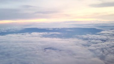 Gün batımında, uçağın penceresinden yayılan canlı bulutlar geniş ufuk boyunca uzanan ılık turuncu ve yumuşak mavi renkleri ile katmanlı gökyüzünü gözler önüne seriyor.