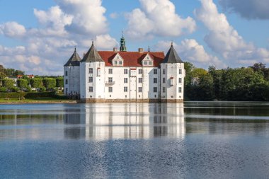 Gluecksburg, Almanya - 29. Ağustos 2025: Güneşli bir günde, mavi gökyüzünde bulutlar olan beyaz şato göle yansıyor