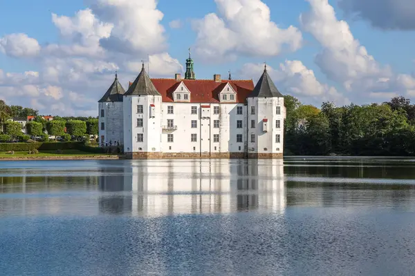 Gluecksburg, Almanya - 29. Ağustos 2025: Güneşli bir günde, mavi gökyüzünde bulutlar olan beyaz şato göle yansıyor
