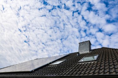 Fotovoltaik paneller, tavan pencereleri ve beyaz bulutlu mavi gökyüzüne karşı bir bacanın yer aldığı modern meskun çatı.