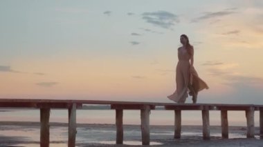 Şık elbiseli bir kadın gün batımında plajda ahşap bir pistte yürüyor.