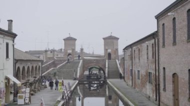 December 2022, Comacchio. People walk by the shore in front of Trepponti monument in a foggy day