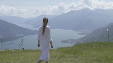 Beyaz giyinmiş bir kadın, İtalya 'nın Como Gölü' nün güzel manzarasının önündeki dağların üzerinde çimenlerde yalınayak yürüyor..