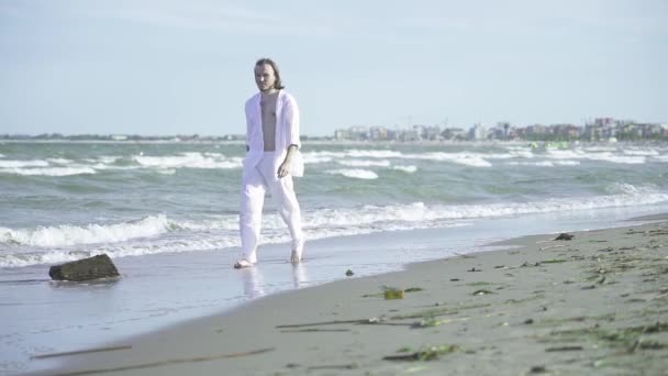 白いスーツの男は風の下で高い波と海によってビーチを歩きます — ストック動画