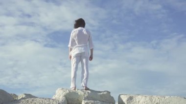 Uzun saçlı adam kollarını rüzgara açıyor, arkadan ve çıplak ayakla taşların üzerinde..