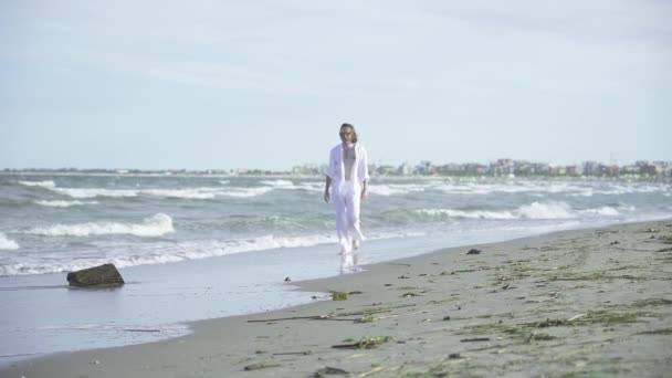 白いシャツとズボンの若い男は 海でビーチでゆっくりと動き 水の中の足で歩きます — ストック動画