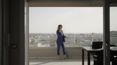 Kadın zarif ve profesyonel giyinmiş, topuklu ayakkabı giyiyor, şehrin yukarısındaki ofis balkonunda telefonla konuşuyor..