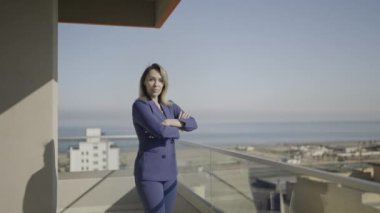 Profesyonel bir kadın, mavi giyinmiş, ellerini katlıyor ve odaya bakıyor, denize bakan binanın balkonuna..
