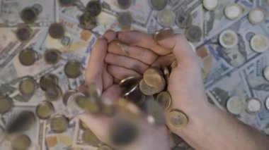 Avro sikkeleri yukarıdan avuçlara yavaş çekimde düşer. Arka planda dağılmış dolar banknotları görülebilir.