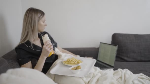 女性は膝の上にラップトップと彼女の手に食べ物のトレイを持つソファーに座っています 彼女の腕は 彼女の指がコンピュータをタップしながら噛むために到達するように伸びています — ストック動画