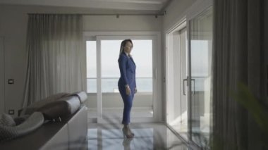 Ciddi bir kadın, mavi elbiseli ve topuklu ayakkabılı, evdeki pencereden içeri bakıyor.