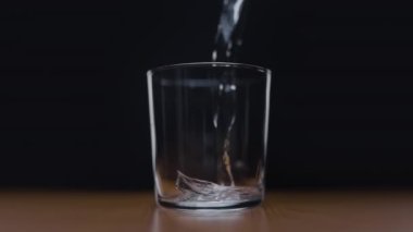 Su bir bardağa dökülüyor, siyah bir duvara ve ahşap bir masaya çarpıyor. Yavaş çekim, suyun dinamik hareketini ve etkisini vurgular.. 
