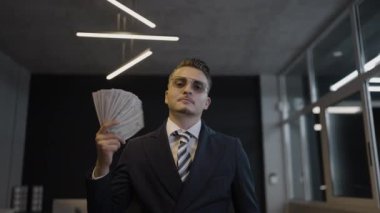 Kendine güvenen bir işadamı, gösterişli bir ofis ortamında özgüvenini sergilerken gururla para yığınları sergiliyor.
