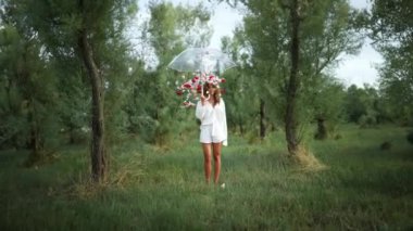 Dalgalı beyaz bir elbise giyen zarif bir kadın neşe içinde gür yeşillikler arasında canlı kırmızı bir şemsiye tutuyor.