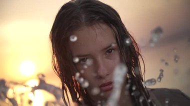 Islak saçlı bir kadın, su damlacıklarının arasından bakar. Parlayan bakışları, dingin bir günbatımının altın rengi tarafından çerçevelenmiş.. 